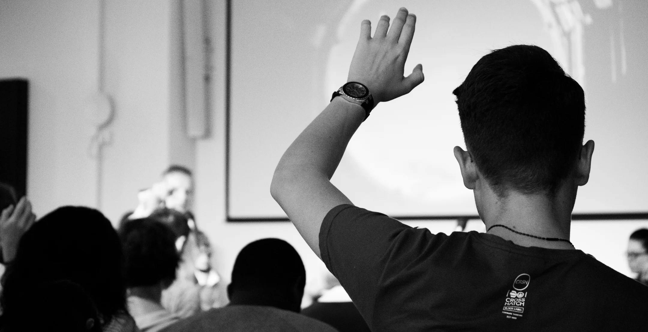 Ein mann rekker opp handa, lerret med presentasjon i bakgrunnen.